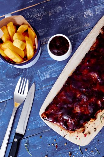 A top view of buffalo ribs, small bowl of sauce, french fries, fork and knife on a wooden blue table