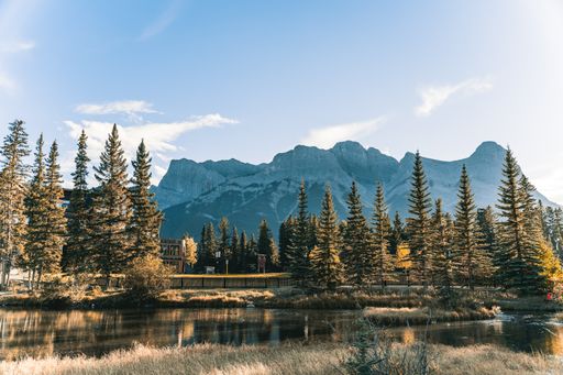 A beautiful wallpaper of a lake and a cottage surrounded by tall fir trees and high snowy mountains in the background