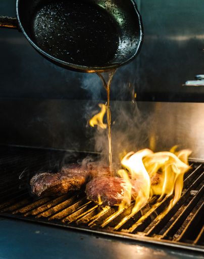 A vertical shot of a pan pouring oil on a grill with burning flames and meat