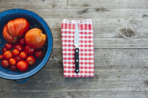 A high angle shot of tomatoes in a blue pot and a knife on a napkin on a wooden surface
