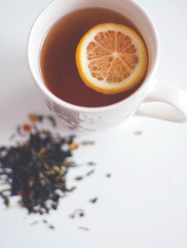 A closeup vertical shot of a white cup of tea with lemon near dried tea leaves
