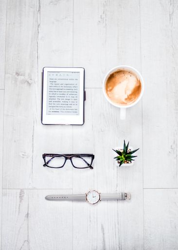 A vertical high angle shot of a tablet, eyeglasses, a watch and a cup of coffee on a wooden table