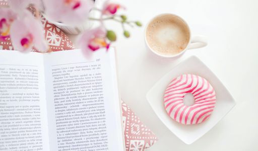 A high angle shot of an opened book near a cup of coffee and a pink donut