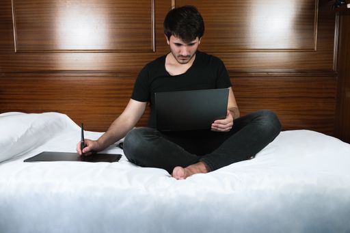 A male working from home with a laptop while sitting in the bed with a wooden background