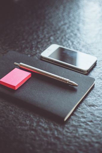 A vertical selective closeup shot of a gray pen and pink sticky notes on a black  notebook near a cellphone