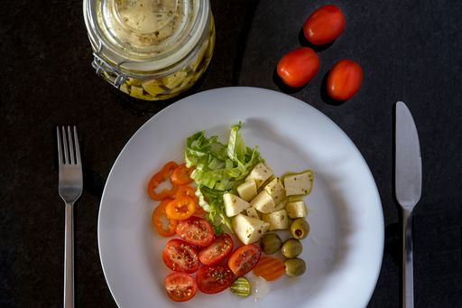 A top view of a salad in a white plate with fork and knife on each side, pickled jar and cherry toma
