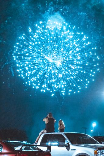 A vertical shot of a father and his daughter sitting on their car and watching blue fireworks