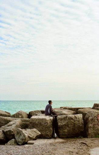 A guy sitting on coastal rocks overlooking the horizon at Kew Balmy Beach, Toronto, Canada