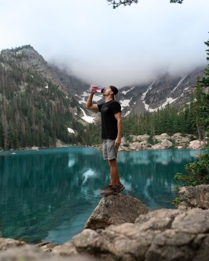 A vertical shot of Caucasian male drinking while standing on a rock by a lake in the mountains