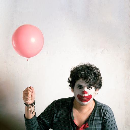 A curly male with a sad clown face holding a pink balloon