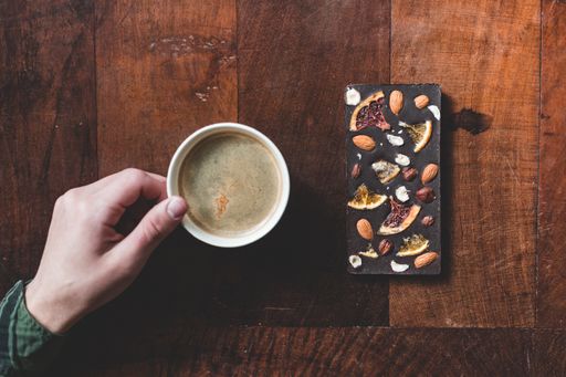 A top view of a man's hand putting a cup of coffee next to a chocolate bar on a wooden table