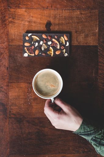 A top view of a man's hand putting a cup of coffee next to a chocolate bar on a wooden table