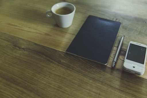 A high angle shot of a notebook with a silver pen, a cup of coffee and white cellphone on wooden table