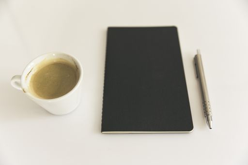 A closeup shot of a black notebook with a pen and a cup of coffee on a white surface