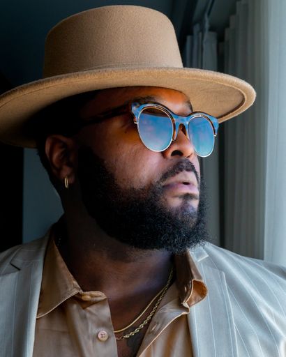 A vertical shot of a male wearing a suit, sunglasses, and a trendy hat, standing near the window