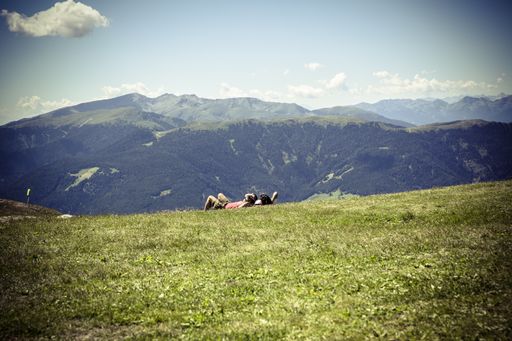 A horizontal shot of two friends lying down on the grass admiring the breathtaking cloudy sky and green mountains