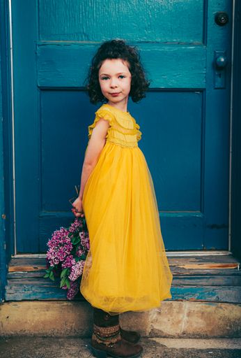 A vertical of a young girl in a bright yellow dress standing at a doorway, holding lilacs