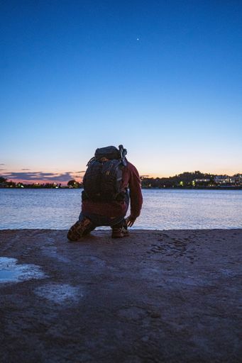A vertical shot of a male with a backpack on knees at the beach at sunset