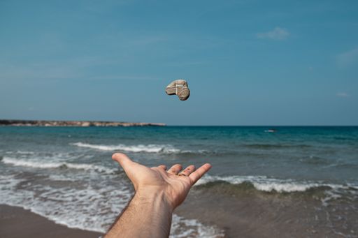 A hand throwing up a rock over a sunny seashore