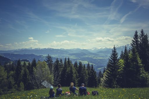 A horizontal shot of four friends sitting on grass enjoying the beautiful view of green trees and mountains under the breathtaking white cloudy sky
