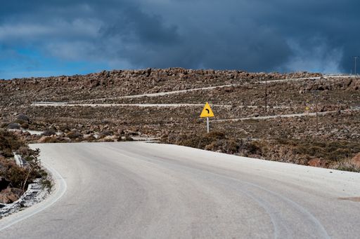 A twisty uphill road with a yellow sign and stormy clouds on the background