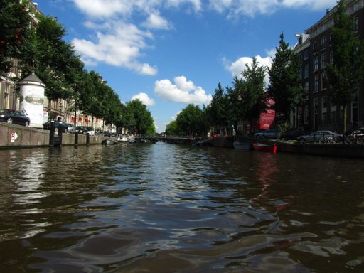 A photo taken from a boat in a canal full of water in Amsterdam, Netherlands, lined with trees and houses on both sides, on a sunny summer day.