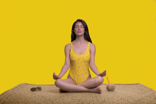 A Hispanic woman sitting in a meditation position next to coconut in a yellow background.