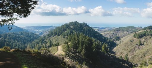 A panoramic shot of mountains covered in greenery under the sunlight and a blue cloudy sky