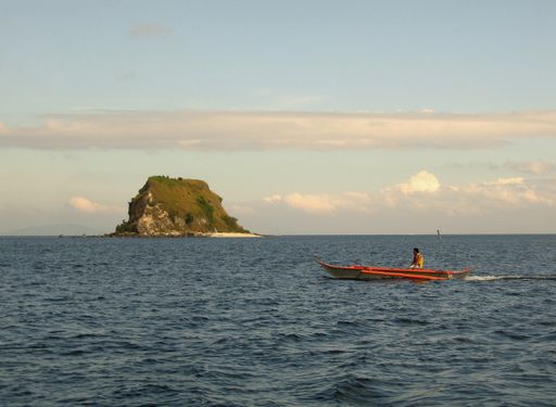 A wide shot of a person riding a canoe on the body of water near a small islet under a blue sky with clouds