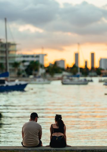 A vertical back view of a couple sitting on a shore and watching golden sunset over a seaside city