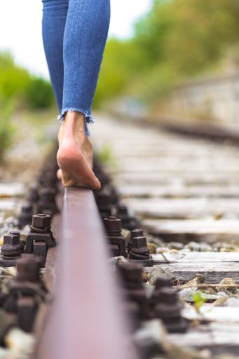 A vertical shot of a female in jeans walking through the train rails barefoot