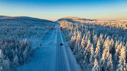Aerial drone view of a car driving in sunlit, snow covered forest of golden hour Lapland