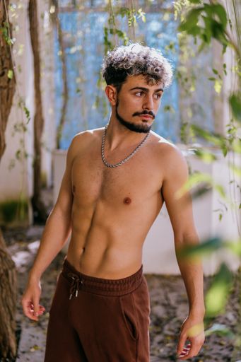 A portrait of an attractive shirtless Spanish man with a beard and hair highlights posing in a garden