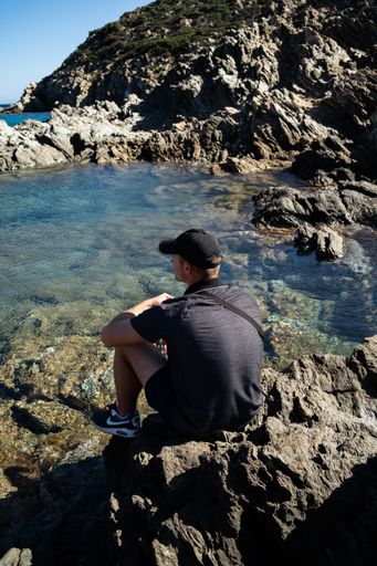 A vertical shot of male sitting on a rock and looking to blue rocky sea in Sardinia