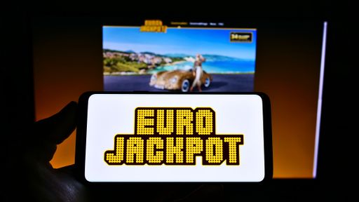 Person holding cellphone with logo of transnational European lottery Eurojackpot on screen in front of business webpage. Focus on phone display.