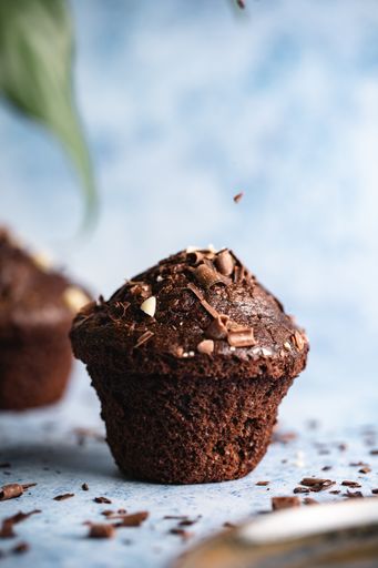 A vertical selective focus shot of chocolate cupcakes on a blue surface with chocolate chips falling