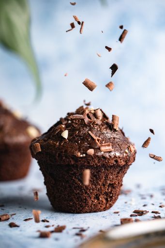 A vertical selective focus shot of chocolate cupcakes on a blue surface with chocolate chips falling