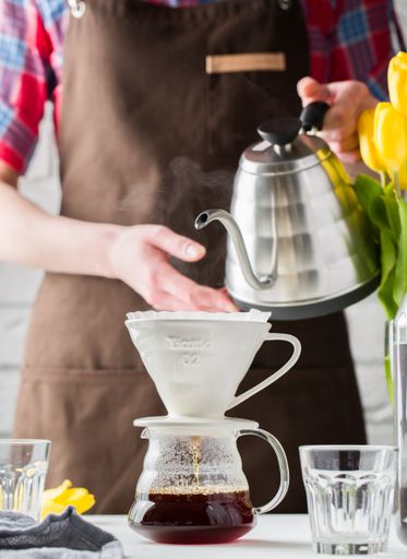 A vertical selective closeup shot of a person holding a kettle pouring hot water into a cup on a glass pitcher for tea