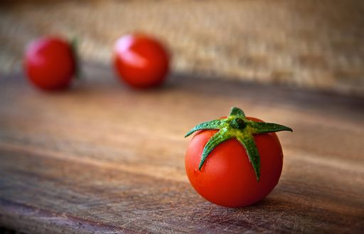 A closeup of a red cherry tomato on a wooden desk with a blurry background of two other tomatoes