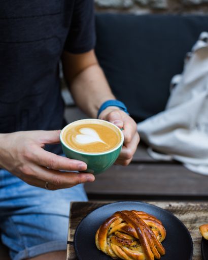 A vertical selective closeup shot of a person holding a cup of latte art coffee near a table with pastry on a plate