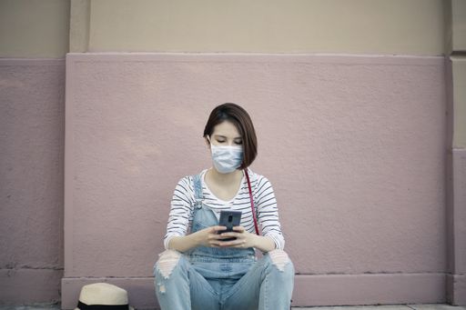 A young female with a facemask sitting on the sidewalk and texting on the phone under the sunlight - COVID-19