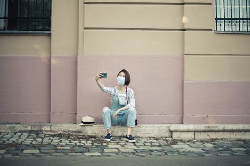 A horizontal shot of a Caucasian female with a face mask sitting on the ground and taking a selfie