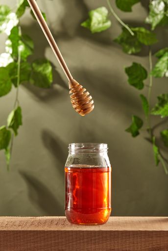 A closeup of honey flowing down the dipper in the glass jar on the green leaves background