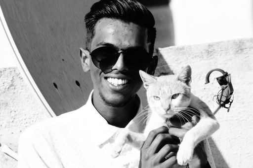 A grayscale of a South Asian man in glasses sitting outdoors with a cat