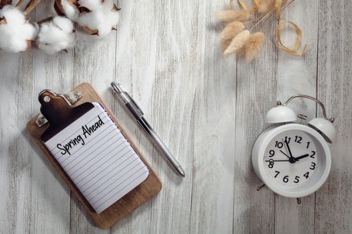 A top view of an alarm clock with a pen and a notepad with "Spring Ahead" text on a wooden table