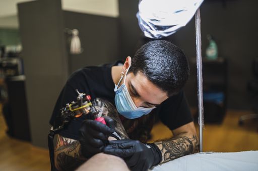 A shot of a male tattoo artist wearing a mask and gloves and tattooing his client's leg