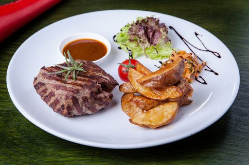 A closeup shot of a delicious lunch with steak, fried potatoes, tomato, salad, and sauce