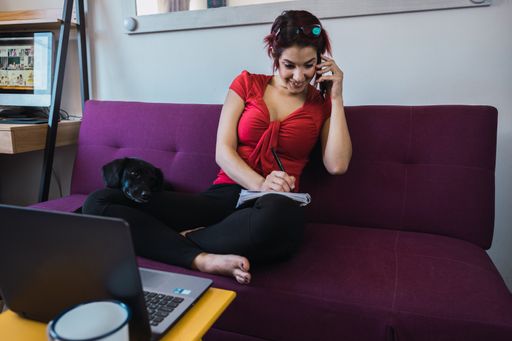 A closeup shot of a young beautiful woman sitting on the sofa with a dog and talking by phone in front of the laptop