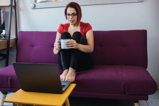A closeup shot of a young beautiful woman holding a cup of tea and sitting on the sofa in front of the laptop