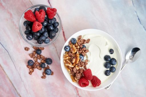 A top view of a bowl with yogurt, raspberries, blueberries, walnuts, and cereals with spoon on a table